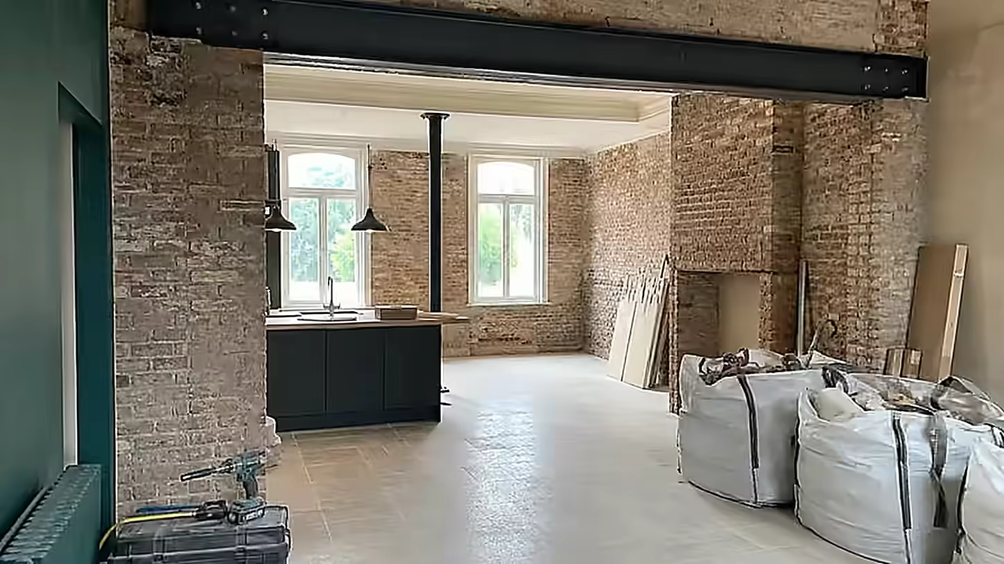 Pièce de vie en cours de chantier, poutre métallique posée, mur de brique conservé et matériaux stockés au sol