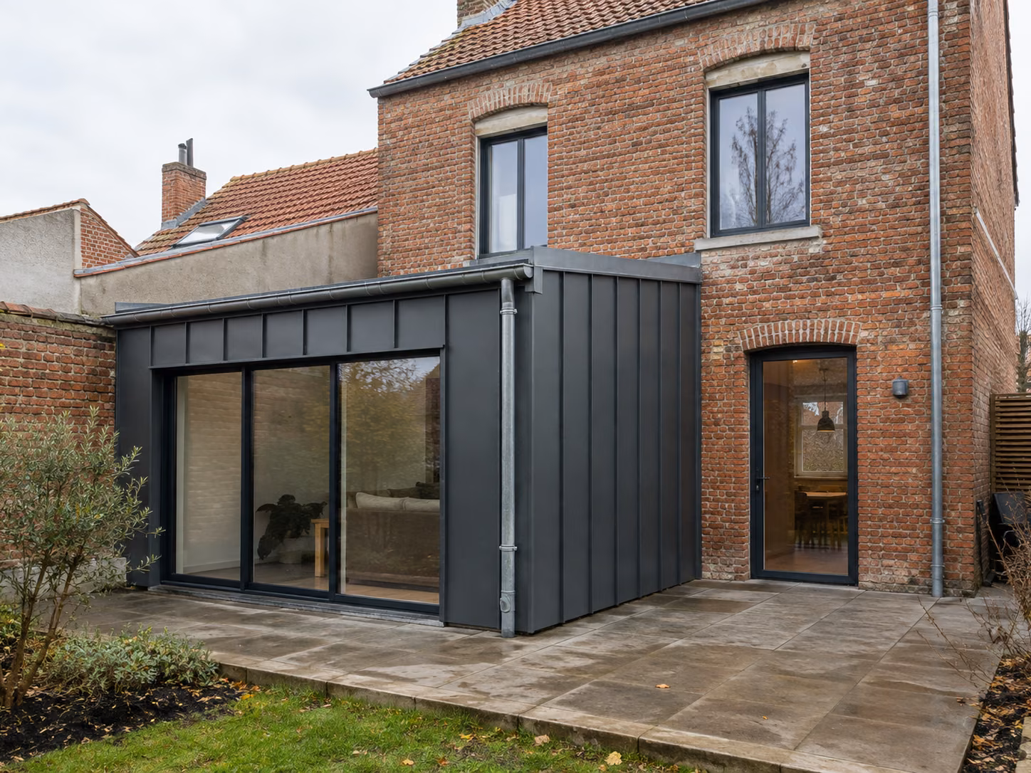 Extension contemporaine bardée de métal sombre greffée à l’arrière d’une maison en brique rouge
