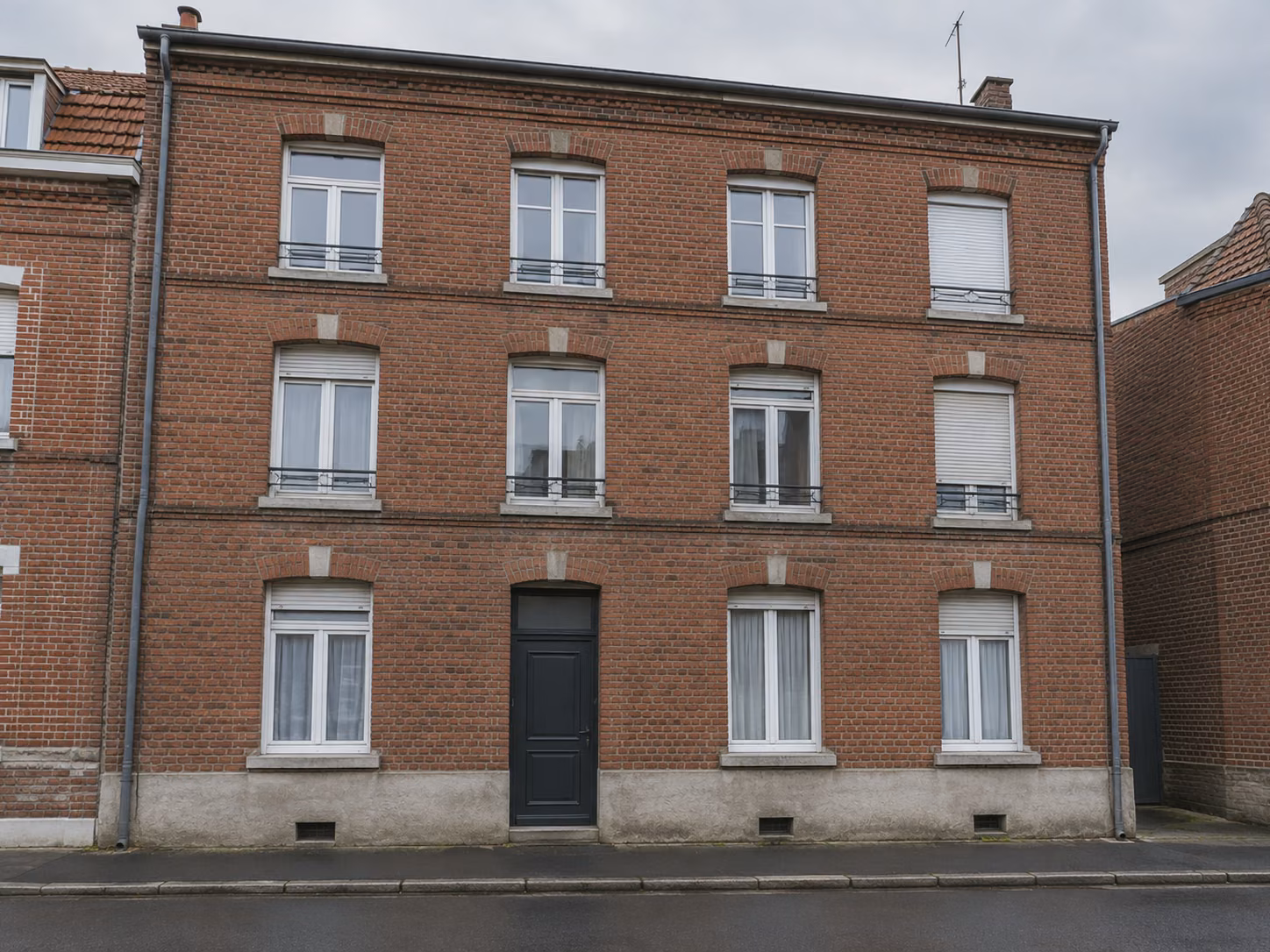 Un immeuble de rapport en brique de trois niveaux vu depuis le trottoir opposé, fenêtres alignées, ciel couvert