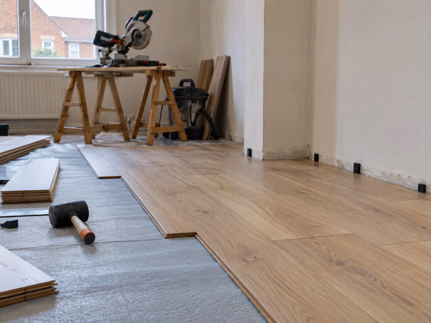 Des lames de parquet stratifié en cours de pose, cales périphériques visibles, découpe à la scie sur tréteaux au fond