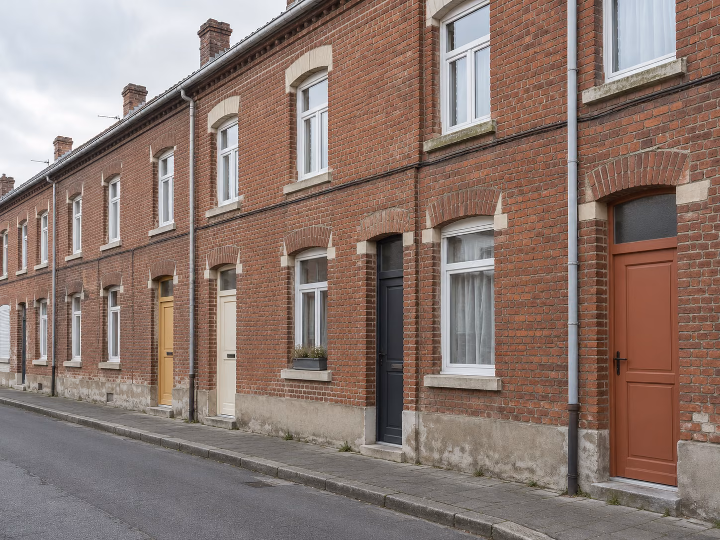 Une rue de corons en brique rouge, façades mitoyennes alignées, portes de couleurs différentes, trottoir étroit, ciel gris lumineux