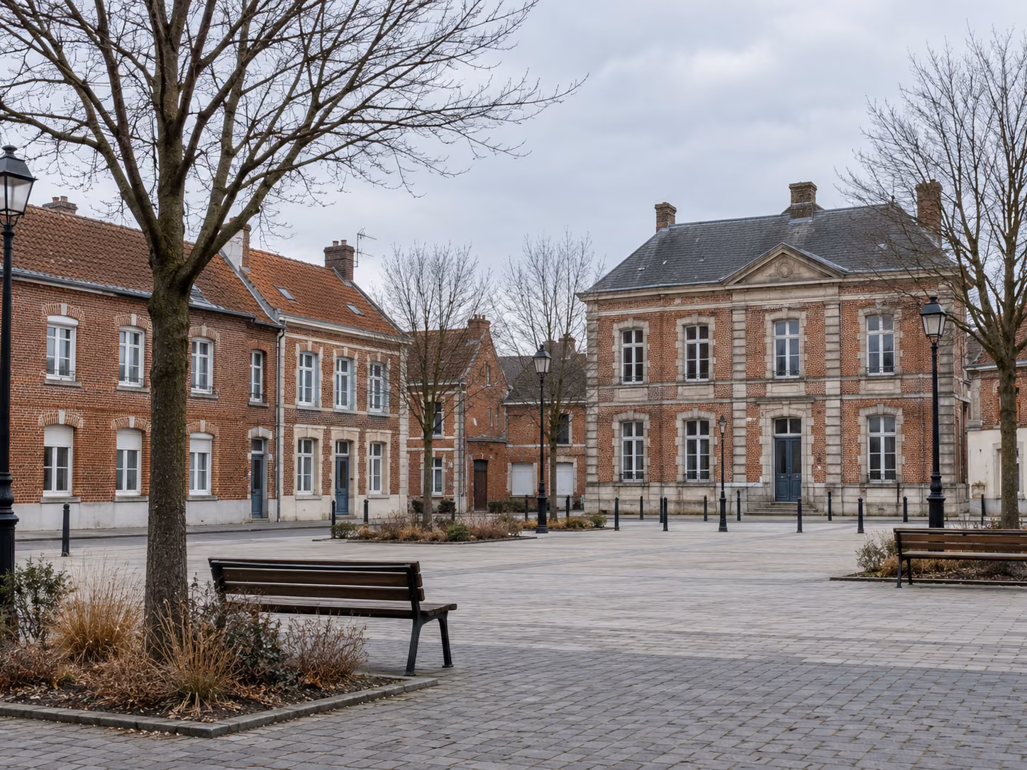 Une place de village bordée de maisons en brique, mobilier urbain simple, arbres sans feuilles, lumière d’hiver