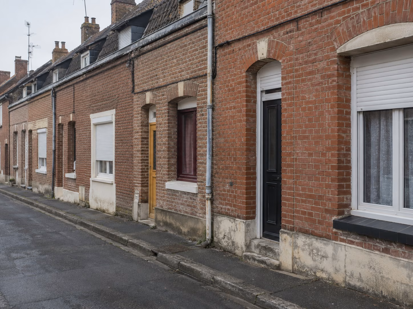 Une rue de corons en brique rouge, façades mitoyennes alignées, portes de couleurs différentes, trottoir étroit, ciel gris lumineux