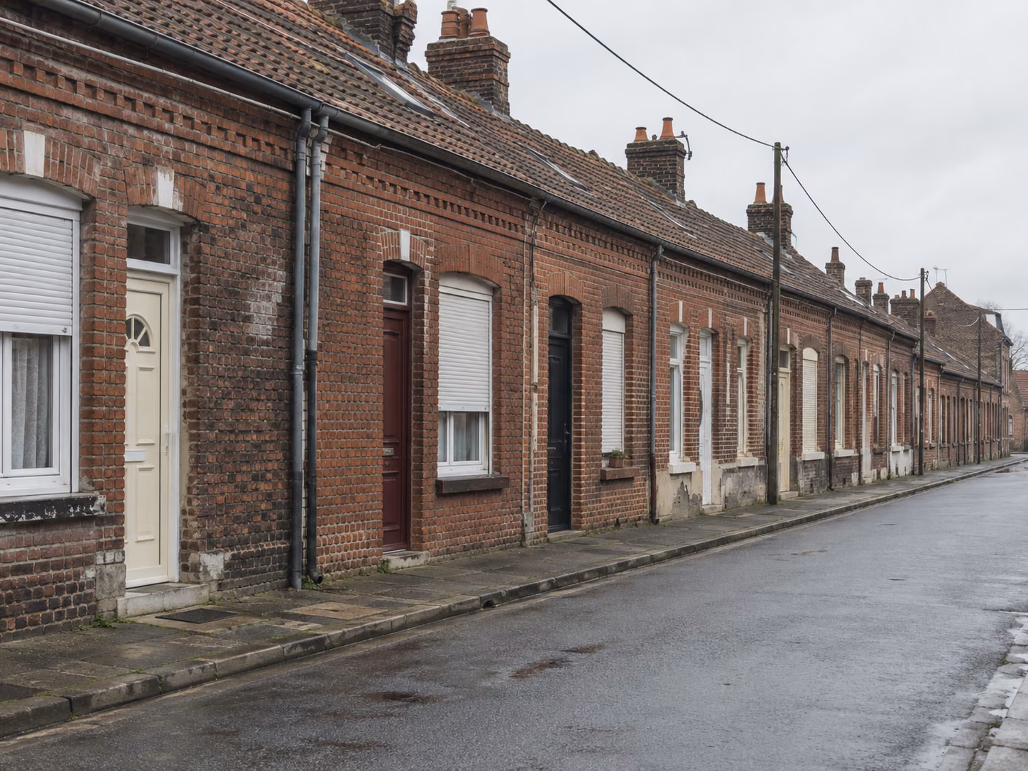Une rue de corons en brique rouge, façades mitoyennes alignées, portes de couleurs différentes, trottoir étroit, ciel gris lumineux