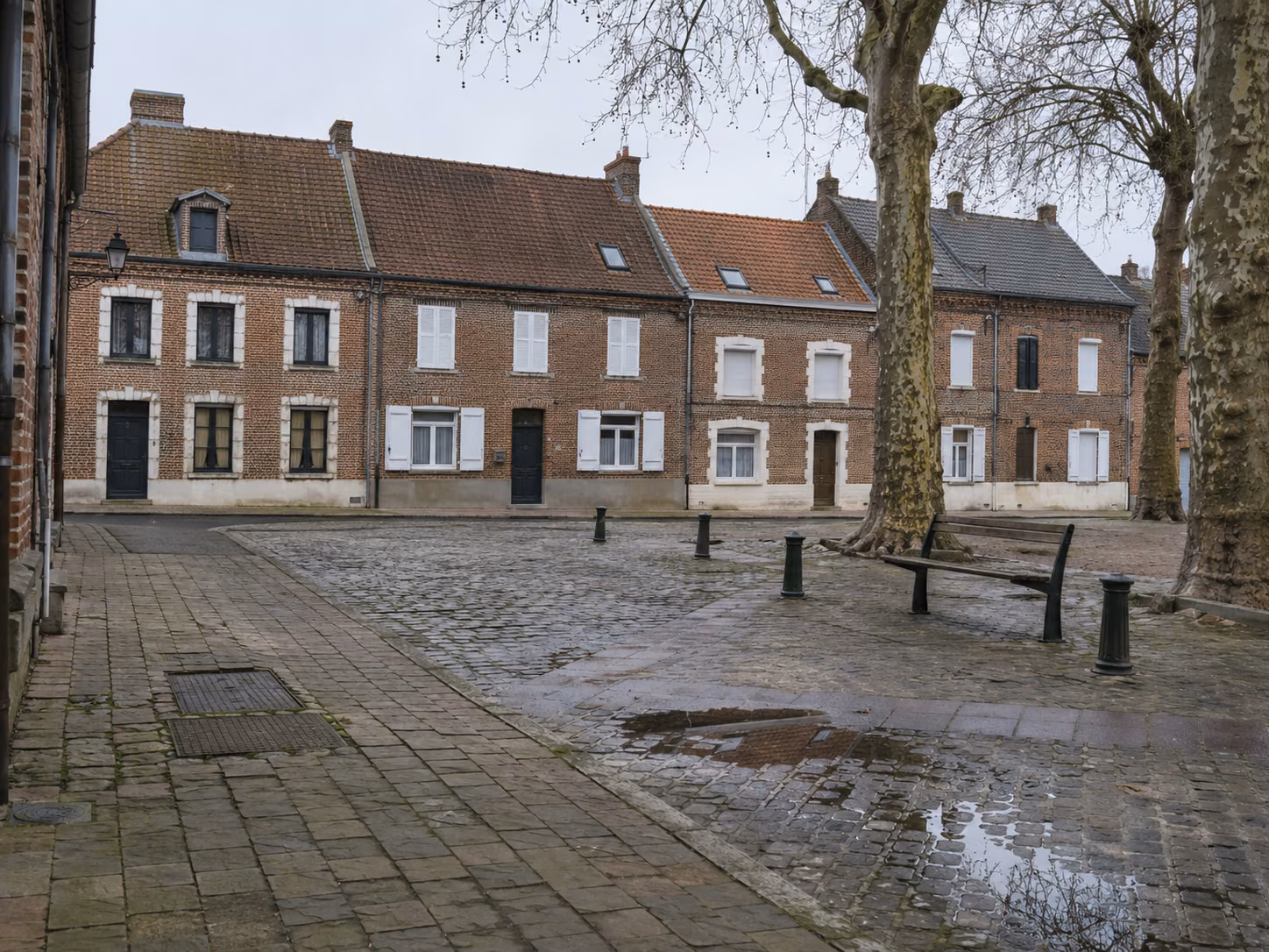 Une place de village bordée de maisons en brique, mobilier urbain simple, arbres sans feuilles, lumière d’hiver