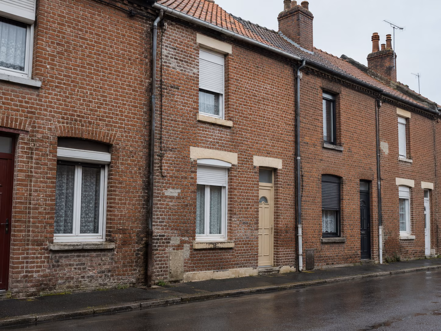 Une rue de corons en brique rouge, façades mitoyennes alignées, portes de couleurs différentes, trottoir étroit, ciel gris lumineux