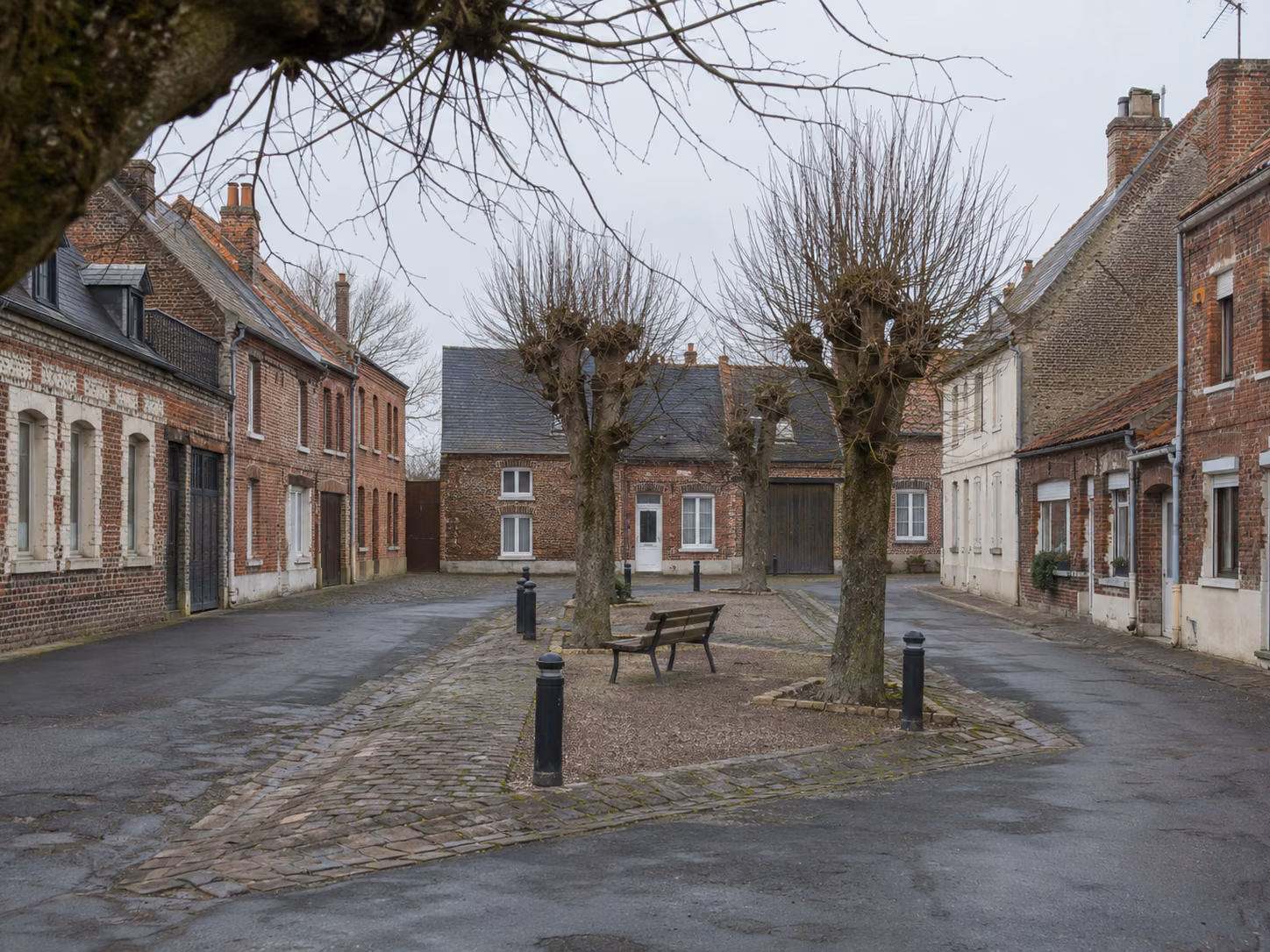 Une place de village bordée de maisons en brique, mobilier urbain simple, arbres sans feuilles, lumière d’hiver
